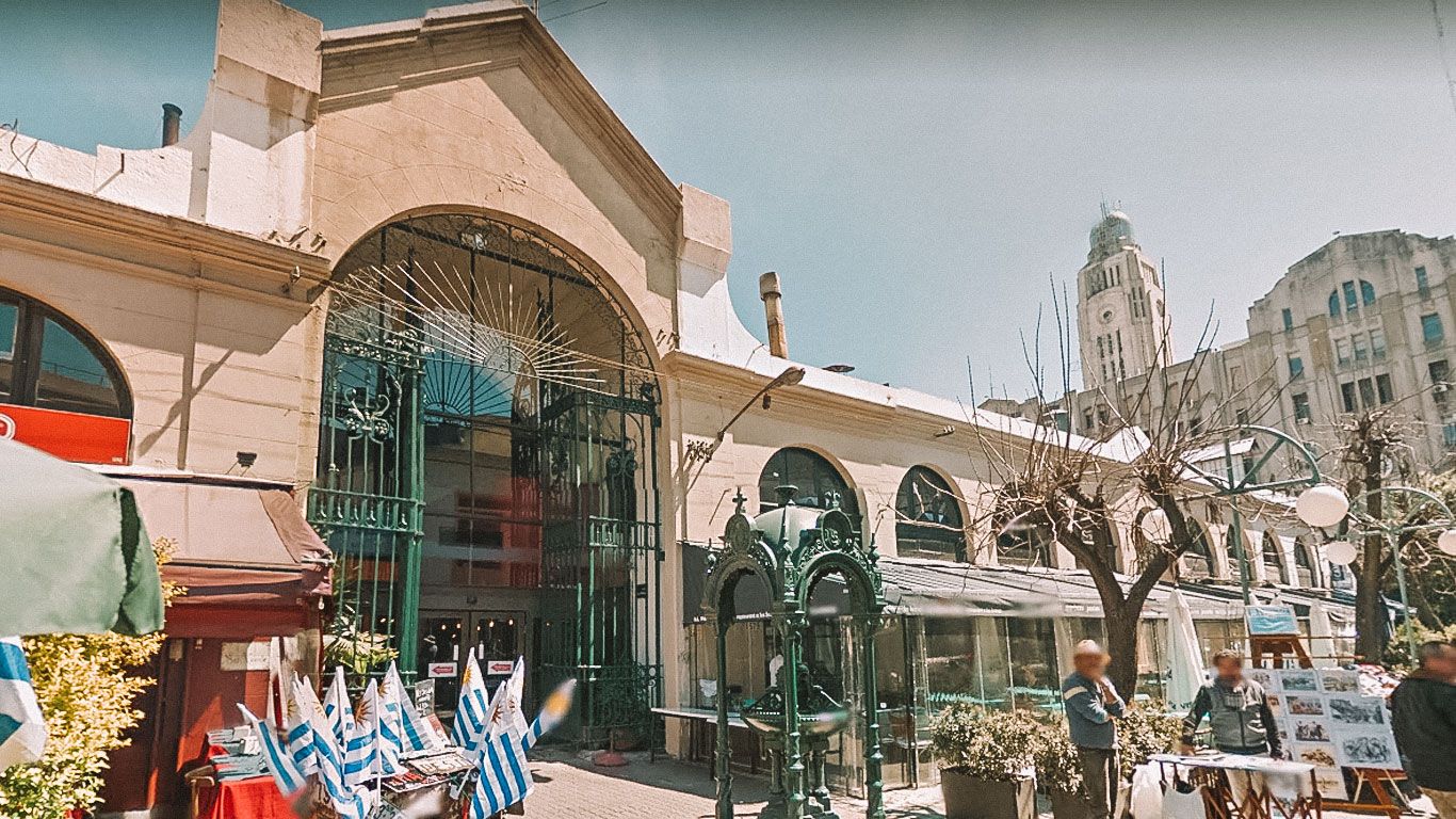 La imagen muestra la entrada principal del Mercado del Puerto, un histórico mercado cubierto con una fachada de piedra y hierro forjado. En el exterior, se ven varias banderas uruguayas y puestos de venta, con personas paseando bajo un cielo despejado y edificios antiguos al fondo.