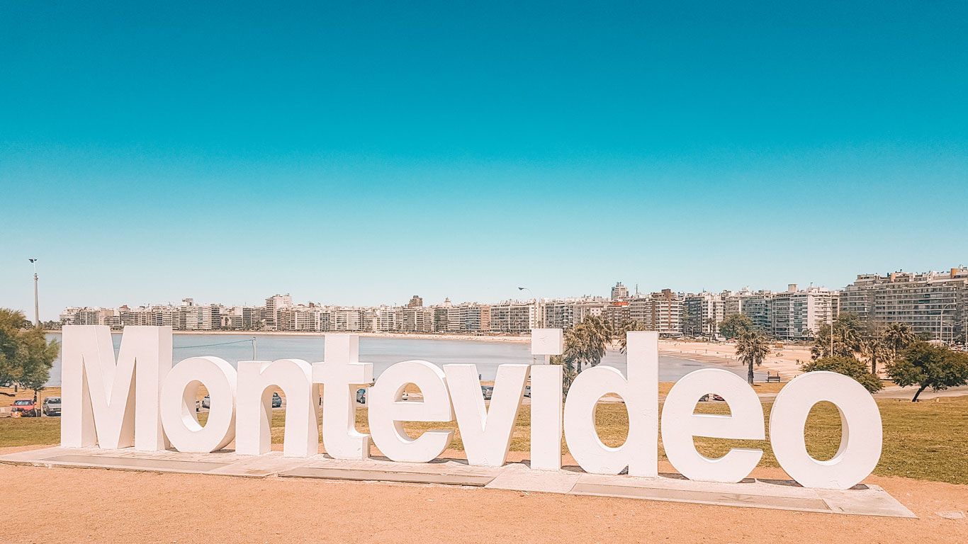 La imagen muestra el icónico cartel de "Montevideo" en la Rambla de Montevideo, con el skyline de la ciudad y la playa al fondo. El cielo es despejado y azul, creando un ambiente perfecto para excursiones en Montevideo. Este punto es popular para tomar fotografías y disfrutar de las vistas panorámicas de la capital uruguaya.