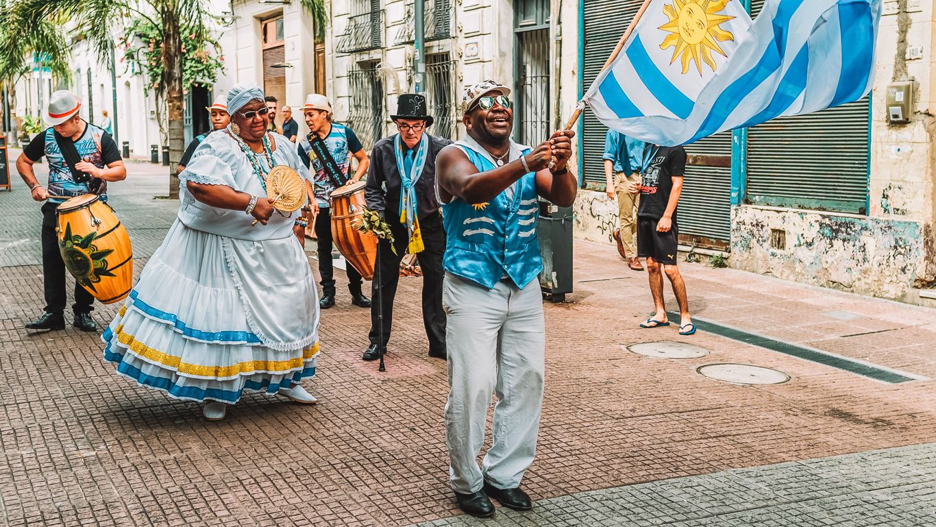 La imagen muestra un animado desfile en Montevideo, Uruguay, con personas vestidas con trajes tradicionales bailando y tocando tambores. Un hombre al frente ondea una bandera de Uruguay con orgullo, mientras una mujer con un vestido blanco y azul sigue el ritmo con un abanico. La escena refleja la rica cultura y tradición festiva de Montevideo.