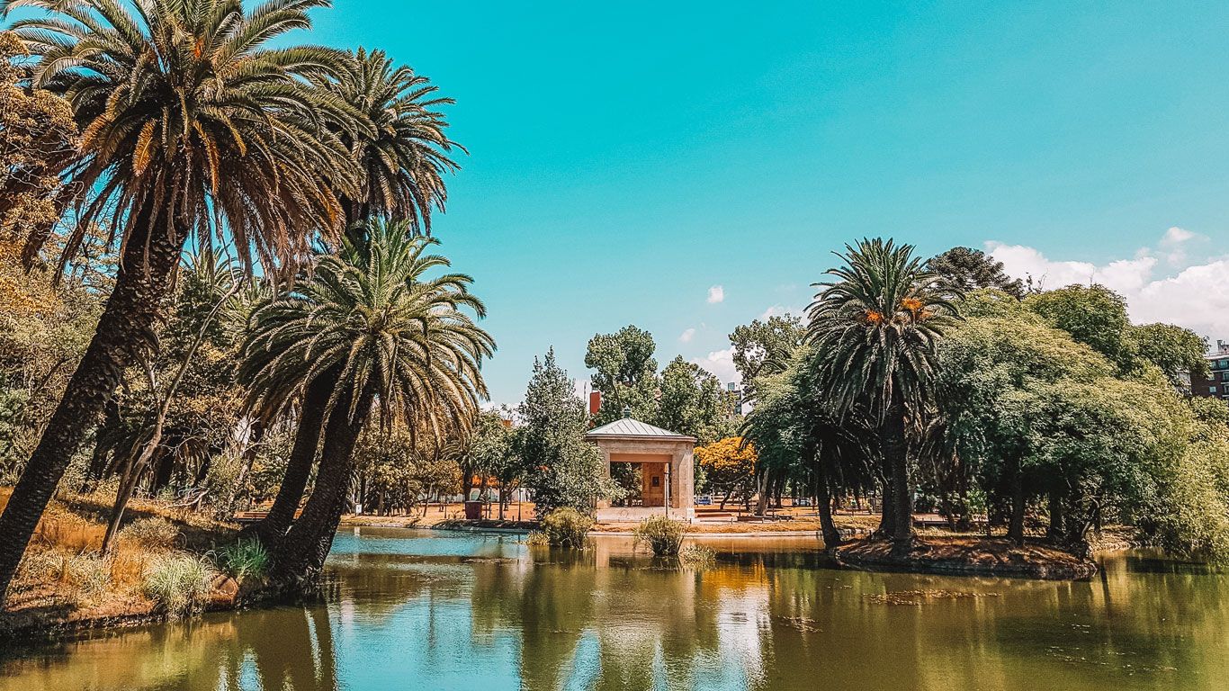 Vista del Parque Rodó en Montevideo, destacando un lago rodeado de palmeras exuberantes y diversa vegetación. En el centro de la imagen, hay una pequeña construcción de estilo clásico a orillas del agua.