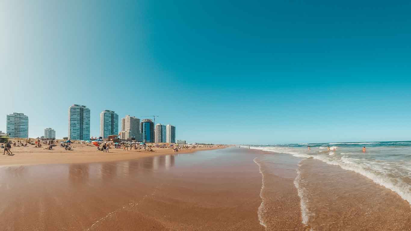 La imagen muestra la popular Playa Brava en Punta del Este, conocida por sus fuertes olas y hermosa arena. En la fotografía se puede observar la línea de costa con su característica arena dorada, las aguas del Atlántico rompiendo suavemente en la orilla y un cielo despejado y soleado que crea un ambiente perfecto para disfrutar del día.