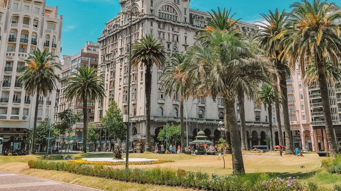 Varias palmeras rodean la Plaza Independencia, un área verde bien cuidada con flores y un pequeño jardín. Al fondo, se puede ver el majestuoso edificio Salvo, acompañado de otros edificios históricos y modernos bajo un cielo despejado.