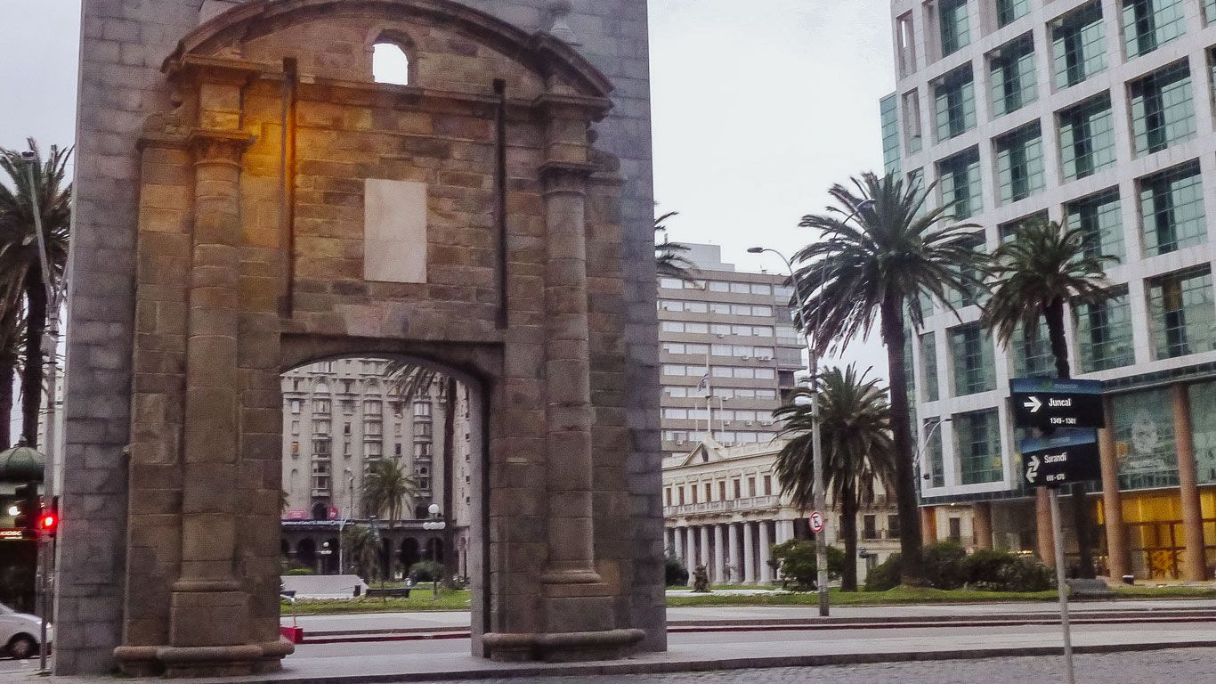 La imagen muestra la histórica Puerta de la Ciudadela, un arco de piedra que antiguamente formaba parte de la muralla de la ciudad. Al fondo, se observan edificios modernos y palmeras en la Plaza Independencia, bajo un cielo nublado.