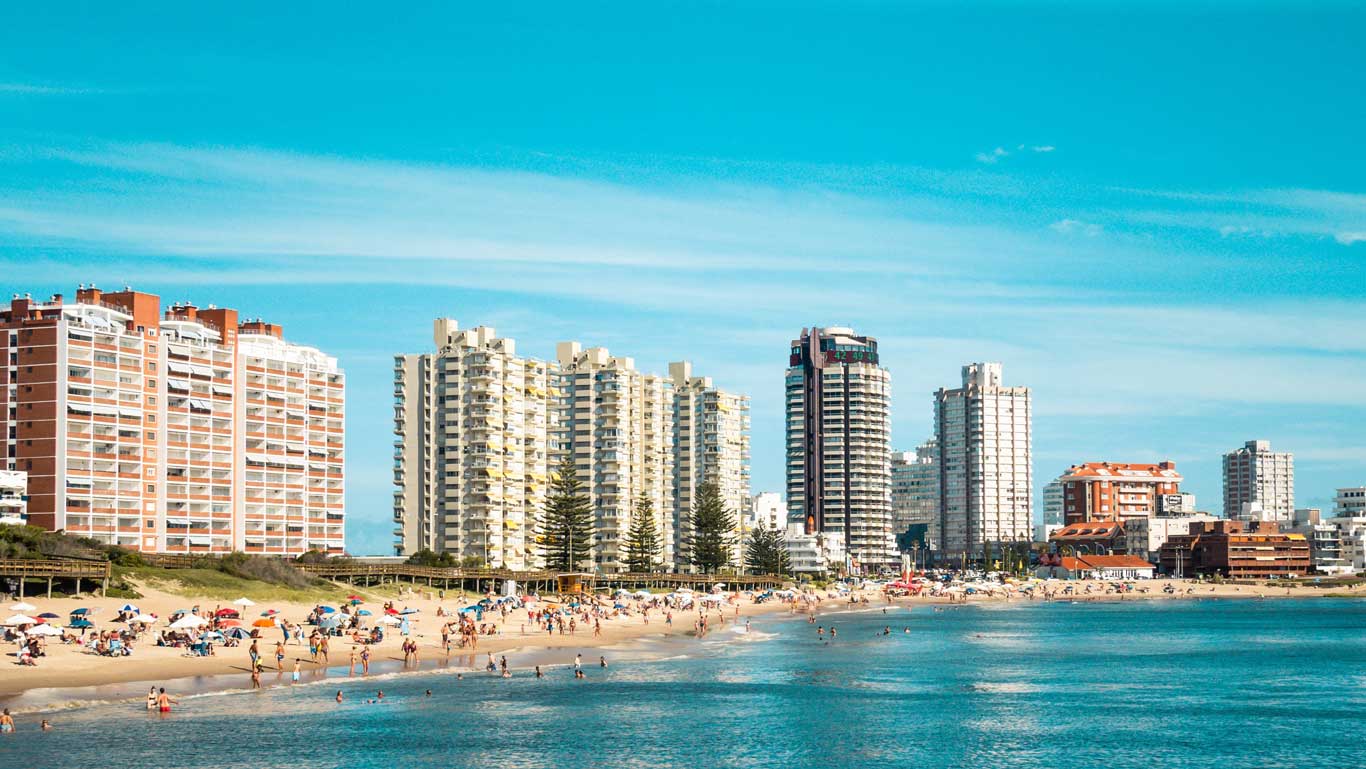 La imagen muestra una playa concurrida en Punta del Este, Uruguay, con edificios altos y modernos en el fondo. Muchas personas disfrutan del sol y del mar, algunas bajo sombrillas. El cielo es azul y despejado, lo que sugiere un día perfecto para actividades al aire libre.