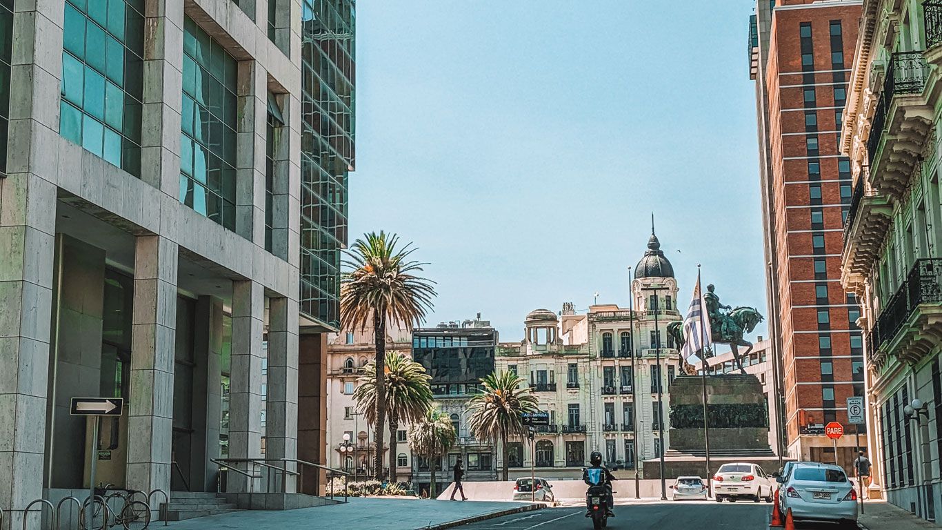 Vista del centro de Montevideo, Uruguay, con edificios modernos y antiguos. En primer plano, una motocicleta en la calle y una estatua ecuestre con la bandera uruguaya ondeando. Palmera y arquitectura histórica se destacan en la escena urbana.