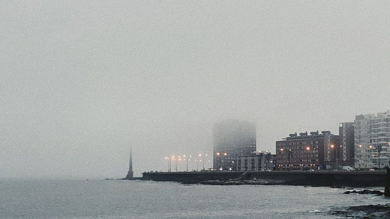 Vista de la rambla de Montevideo en un día nublado, con edificios y luces urbanas a lo largo de la costa. La niebla cubre parcialmente los edificios, creando una atmósfera serena y misteriosa. Este escenario destaca la belleza costera y la vida urbana de Montevideo.