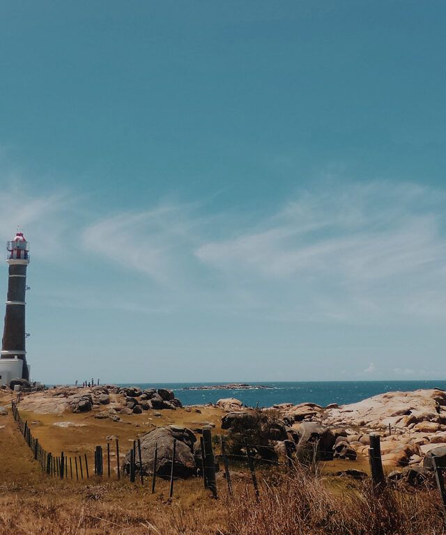 Faro de Cabo Polonio en Uruguay, rodeado de construcciones blancas bajas y un paisaje costero rocoso con pasto seco. Al fondo, el océano Atlántico se extiende bajo un cielo azul con nubes finas, creando una vista serena y natural.