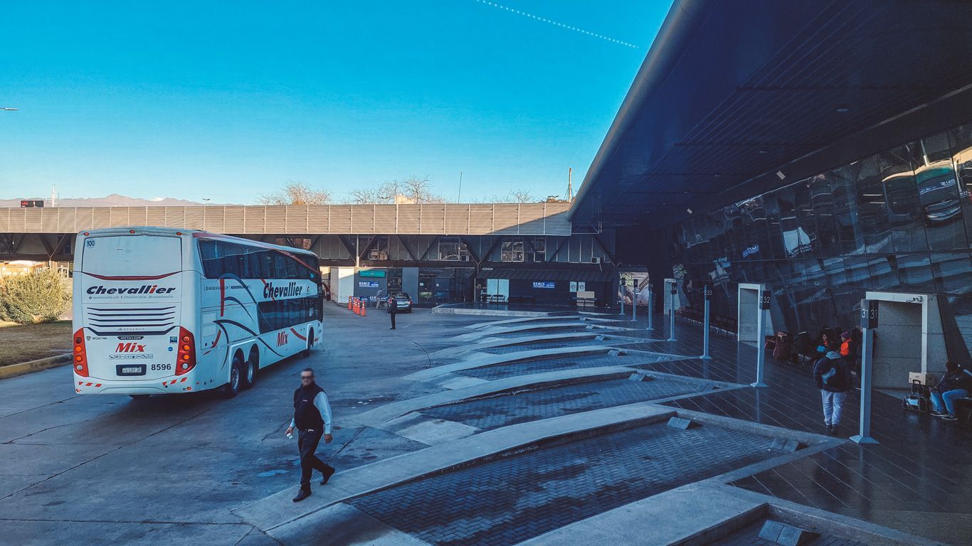 Un autobús de la empresa Chevallier estacionado en una terminal de transporte, con plataformas numeradas y varias personas esperando. El cielo despejado y la estructura moderna de la terminal reflejan un entorno ordenado y bien iluminado. El autobús es una de las formas de hacer el viaje de Buenos Aires a Mendoza.