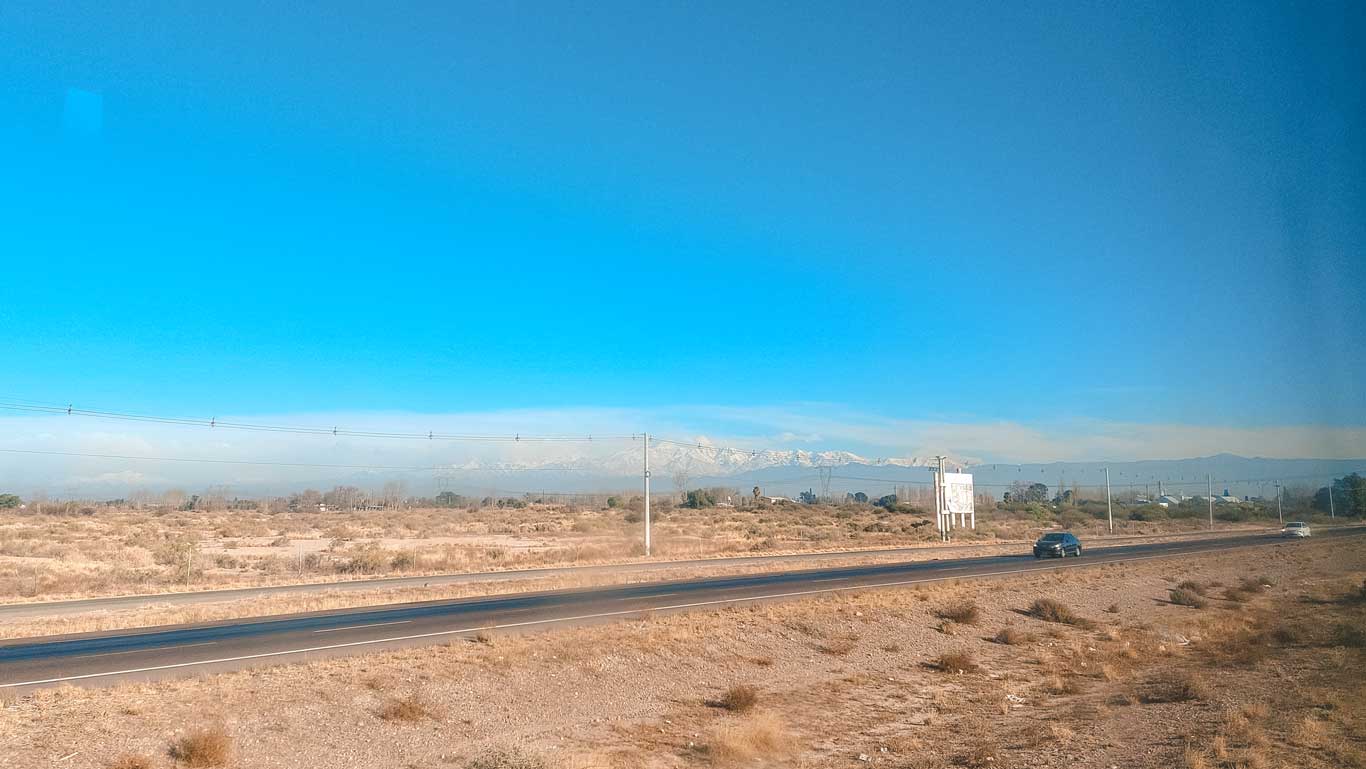 Una carretera desértica rodeada de vegetación seca con un paisaje montañoso al fondo, destacando picos nevados bajo un cielo despejado. Algunos vehículos transitan por la carretera, mientras postes y cables eléctricos cruzan el horizonte.