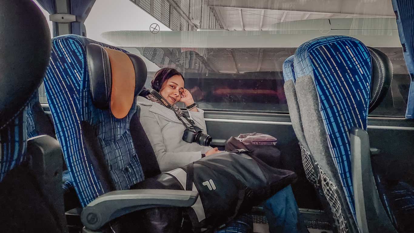 Mujer con una ligera sonrisa en el rostro, sentada en un asiento del autobús de Montevideo a Colonia del Sacramento, junto a una mochila y con una cámara colgada al cuello.