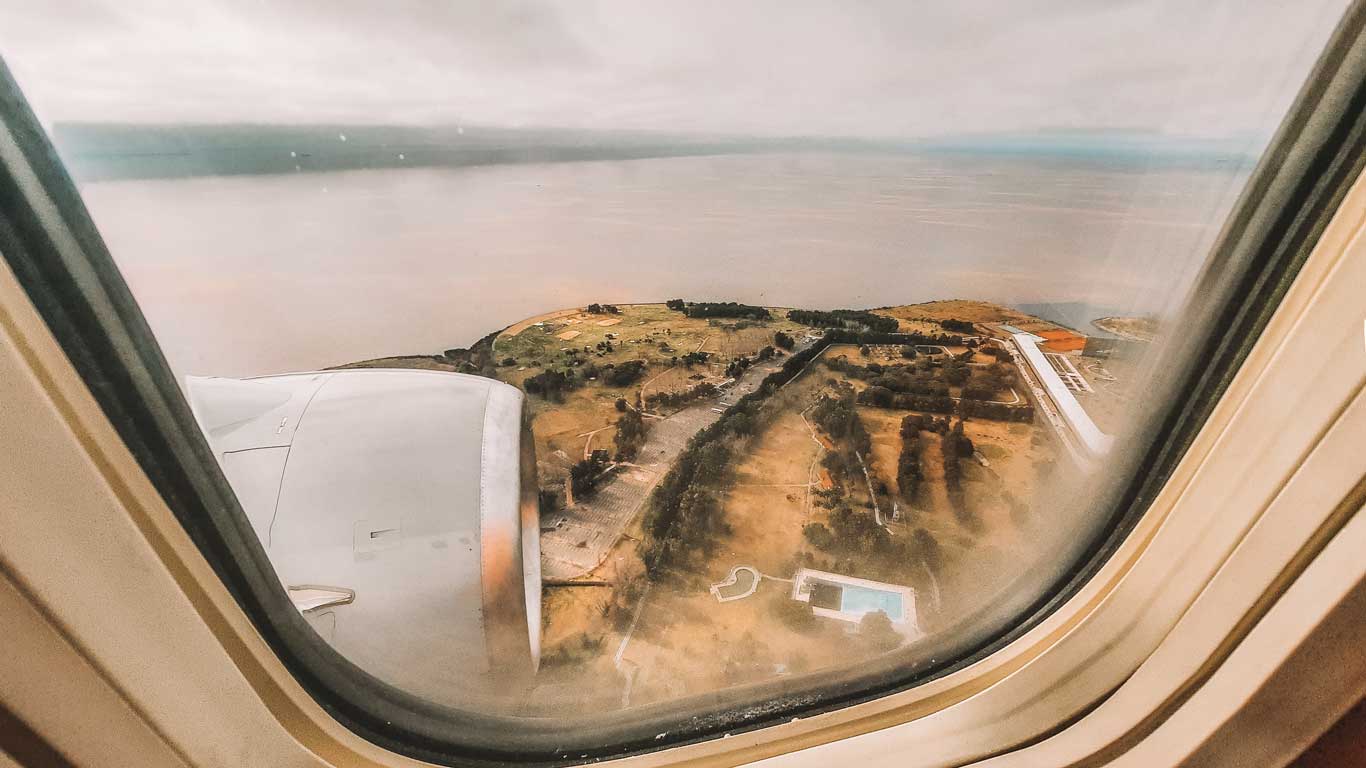 Un paisaje visto desde la ventana de un avión, mostrando un motor en primer plano y un área costera con vegetación, estructuras y una piscina visible en la parte inferior derecha. El cielo está nublado, con el agua extendiéndose en el horizonte.