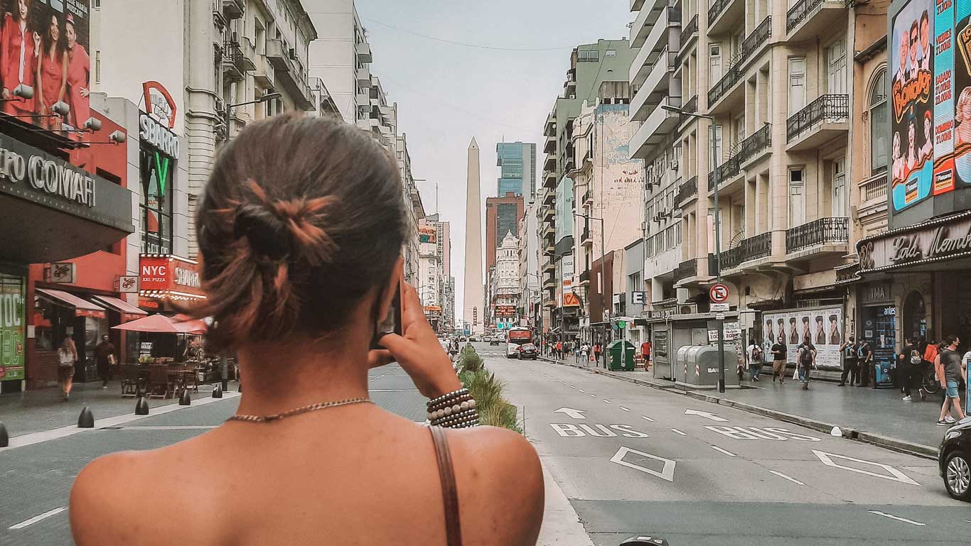 La imagen muestra una calle céntrica de Buenos Aires, Argentina, con el icónico Obelisco al fondo. Una mujer de espaldas toma una foto del monumento, rodeada de edificios altos y carteles publicitarios. La escena captura la vibrante vida urbana de Buenos Aires.