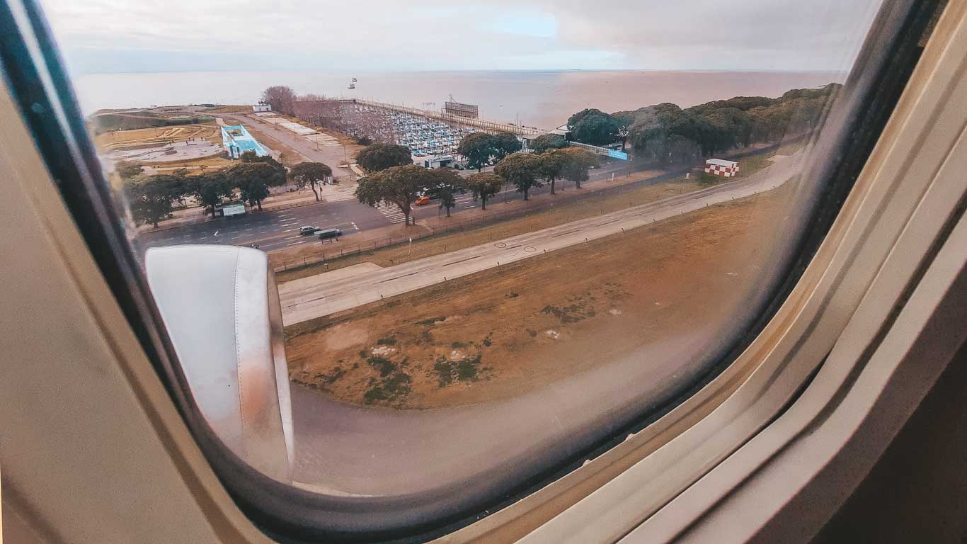 La imagen muestra una vista aérea tomada desde un avión que sobrevuela una zona costera, posiblemente en aproximación a un aeropuerto. Se pueden ver una carretera, árboles, edificios y el océano en el fondo. Esta vista sugiere una opción de como ir de Buenos Aires a Montevideo en avión.