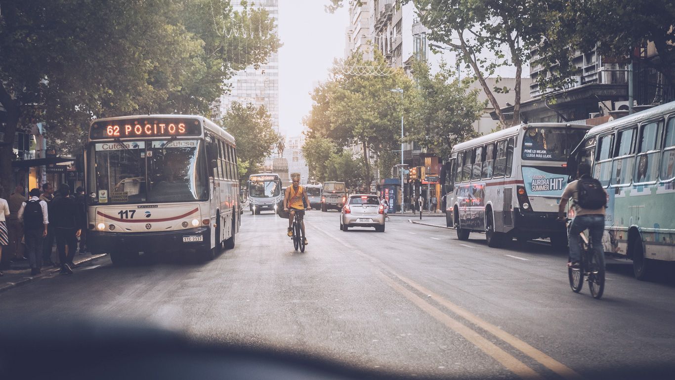 La imagen muestra una calle transitada de Montevideo, Uruguay, con un autobús de la línea 62 con destino a Pocitos en primer plano. También se ven ciclistas, coches y peatones, además de árboles y edificios que bordean la calle. El ambiente urbano es animado y concurrido, destacando el dinamismo de la ciudad.