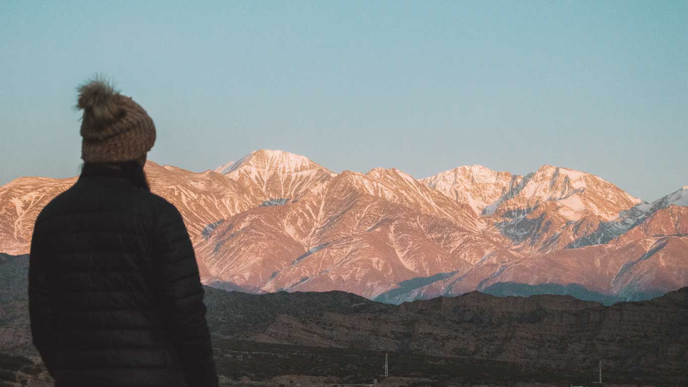 Una figura solitaria con atuendo de invierno se encuentra en contemplación, mirando los picos besados por el sol de los Andes en Mendoza, con los tonos anaranjados del amanecer o el atardecer pintando las montañas nevadas.