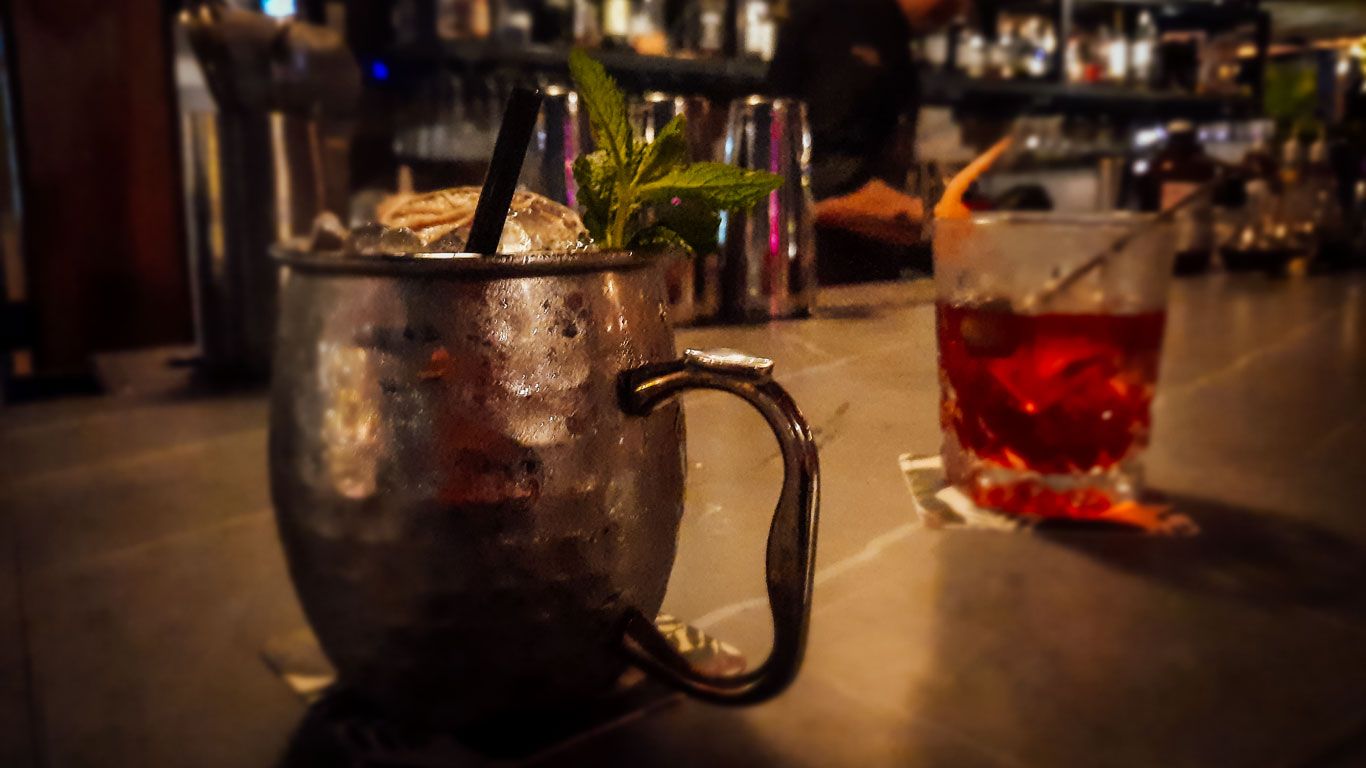 En esta imagen tomada en un bar, se observan dos bebidas en una barra: un cóctel servido en una taza metálica con hielo triturado y hojas de menta, y otro cóctel rojo en un vaso de cristal con una cáscara de naranja. El fondo está desenfocado, mostrando botellas y utensilios de bar, lo que sugiere un ambiente acogedor y sofisticado.