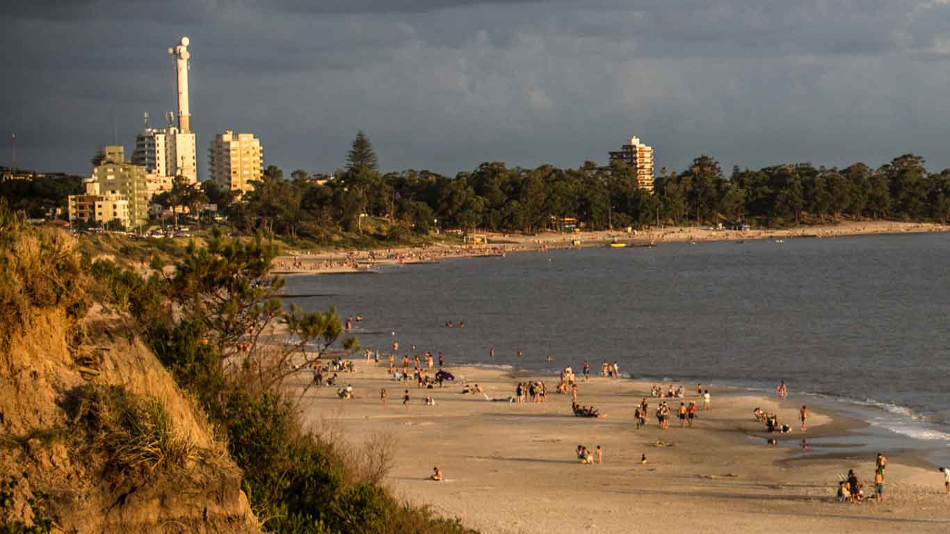 La imagen muestra la playa de Piriápolis en Uruguay, con varias personas disfrutando del día. En el fondo, se observan edificios y una torre de telecomunicaciones, rodeados de árboles y vegetación. El cielo está nublado, pero el ambiente en la playa es animado y concurrido.