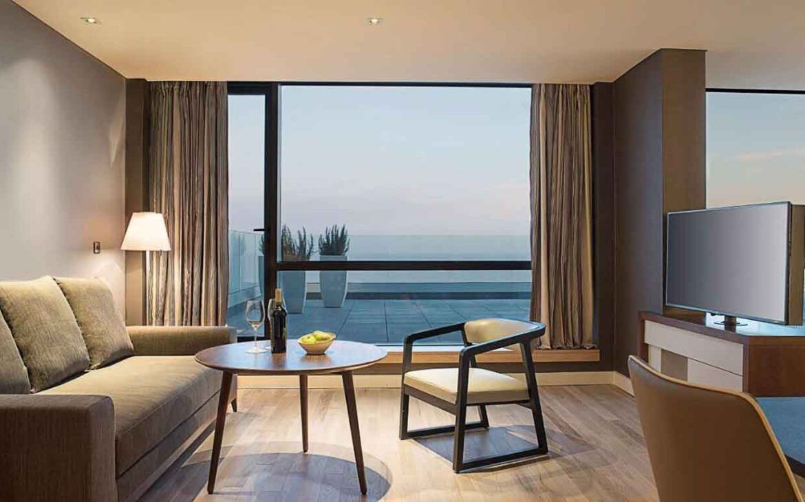 La imagen muestra una elegante sala de estar en una suite del Hyatt Centric Montevideo. La habitación está decorada con muebles modernos y minimalistas, incluyendo un sofá, una mesa de café y una silla. Los grandes ventanales ofrecen una vista espectacular del océano, creando un ambiente relajante y sofisticado.