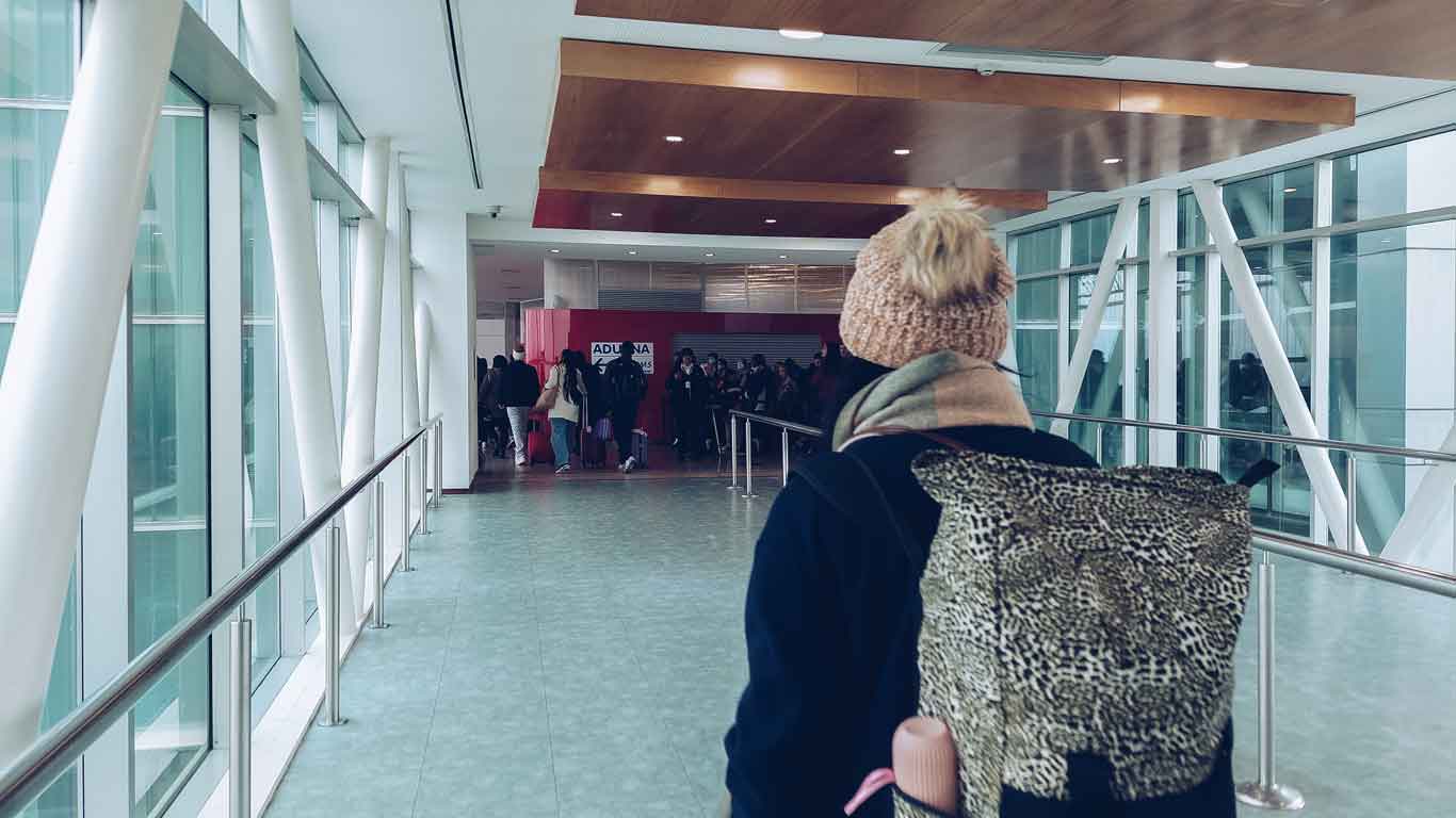 La imagen muestra a una persona de espaldas con una gorra de punto y una mochila con estampado de leopardo, observando a un grupo de personas que parecen estar en una fila frente a aduana uruguaya. La escena ocurre en un interior amplio y moderno, con paredes blancas y ventanas de piso a techo que dejan entrar luz natural, y un techo de madera que añade un toque cálido al ambiente.