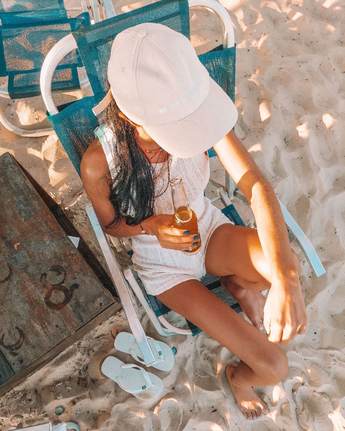 Mujer relajándose en una silla de playa en las arenosas costas de Barra da Tijuca, Río de Janeiro, disfrutando de una bebida fría, con un par de chanclas blancas en primer plano.