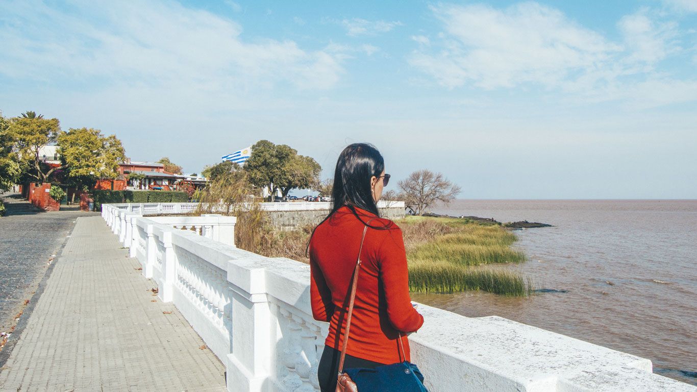 La imagen muestra a una mujer de espaldas, contemplando el paisaje del Río de la Plata desde un paseo en Colonia del Sacramento, Uruguay. Ella lleva una chaqueta roja y una cartera cruzada marrón. La escena incluye una valla blanca, abundante vegetación verde y el cielo despejado, con un fondo de edificaciones y una bandera uruguaya.
