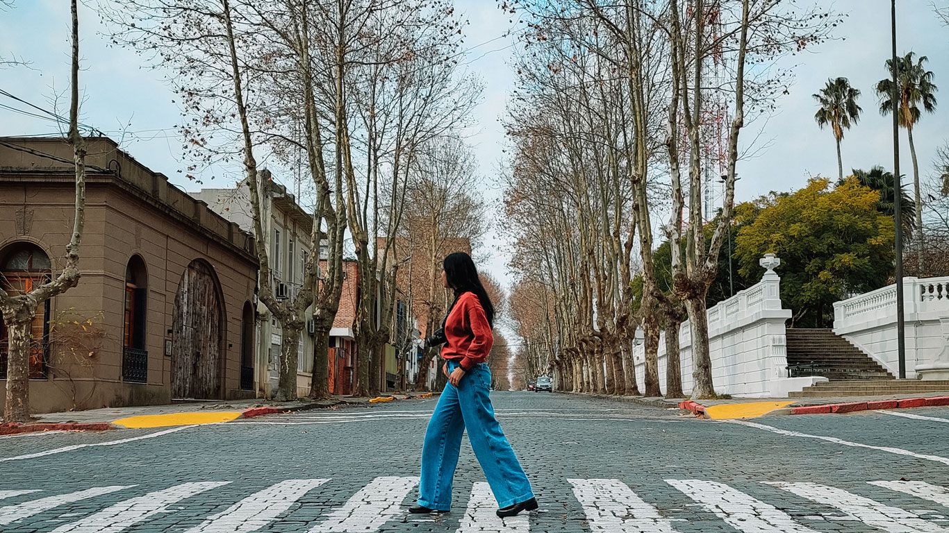 La imagen muestra a una mujer caminando por una calle empedrada en Colonia del Sacramento. Ella lleva una chaqueta roja y jeans azules. A ambos lados de la calle, hay filas de árboles altos despojados de hojas, sugiriendo que es otoño o invierno. Al fondo, se observan algunos edificios antiguos y escaleras que conducen a un parque o plaza, creando una atmósfera histórica y tranquila.