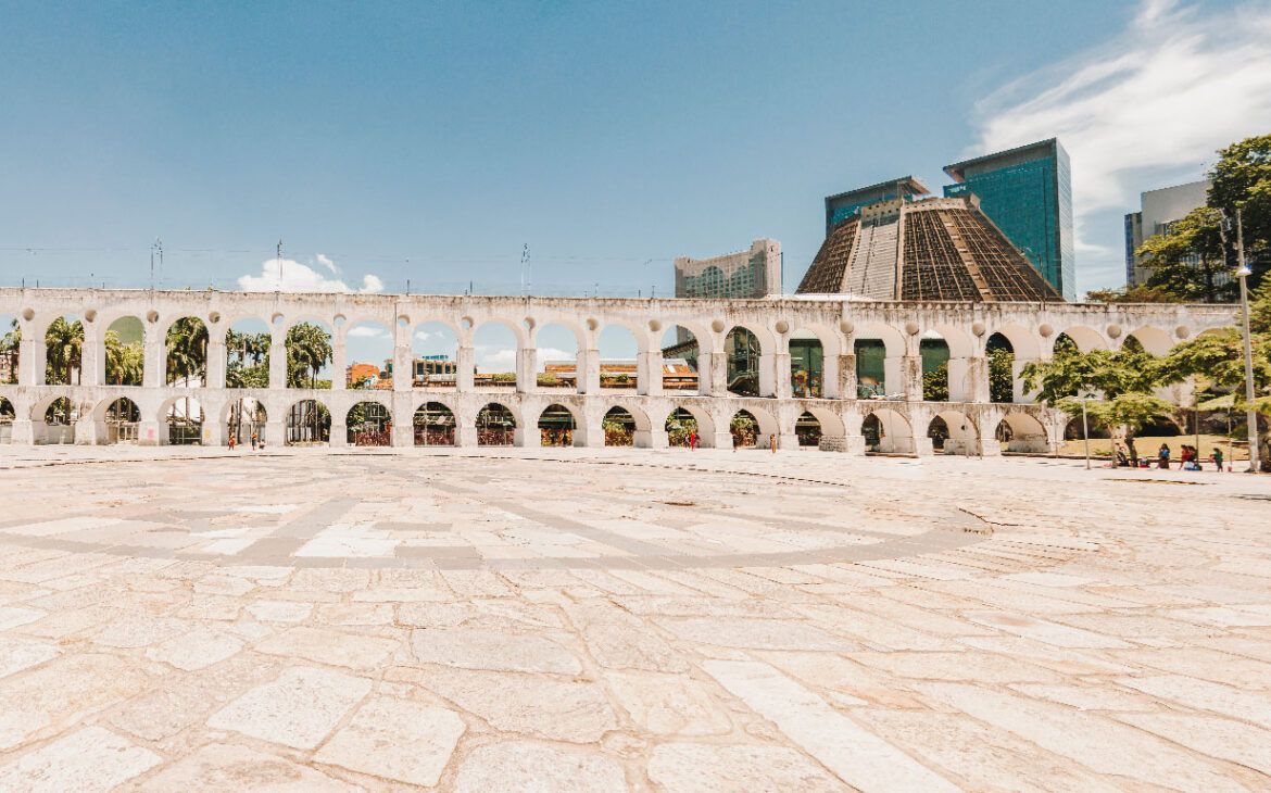 El histórico acueducto Arcos da Lapa en Río de Janeiro se destaca prominentemente contra un cielo azul brillante, con su estructura colonial contrastando con edificios modernos en el fondo, un punto de referencia popular en el corazón de la ciudad.