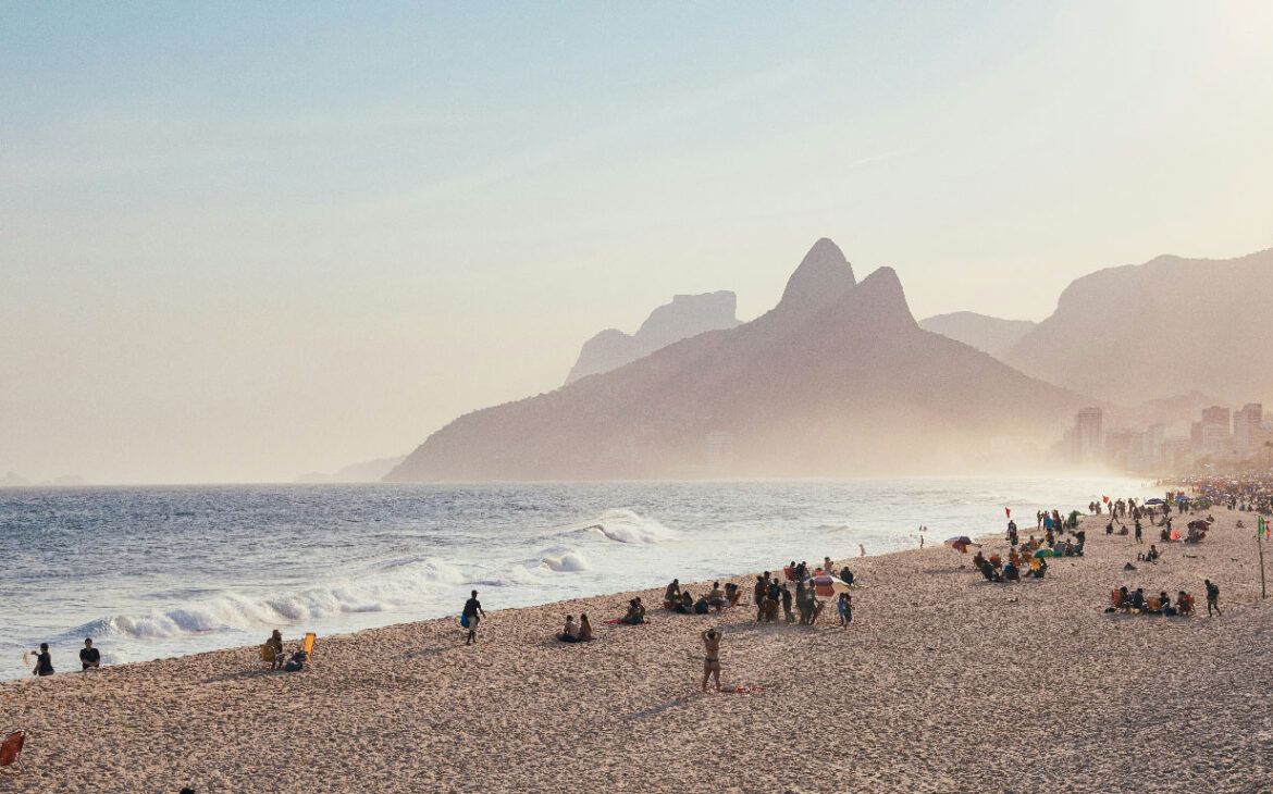 La hora dorada proyecta un resplandor cálido sobre la playa de Leblon en Río de Janeiro, con personas disfrutando de la playa arenosa y el distintivo monte Dos Hermanos (Morro Dois Irmãos) elevándose majestuosamente al fondo.