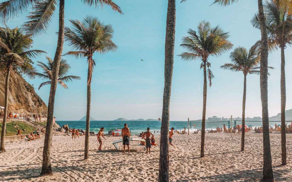 Personas disfrutando de un día soleado en la playa de Leme en Río de Janeiro, jugando vóleibol de playa entre altas palmeras, con las suaves olas del Océano Atlántico y la pintoresca ladera al fondo.