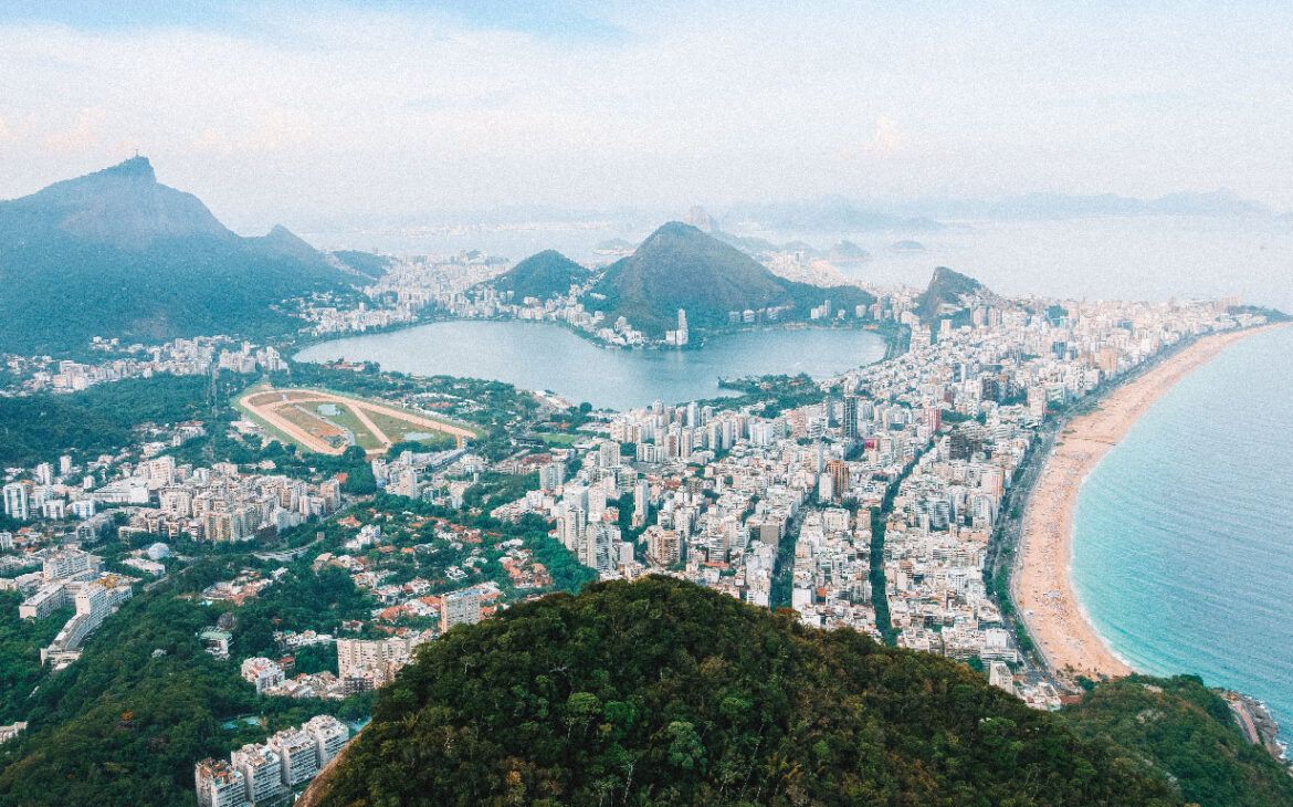 Vista aérea del barrio de Vidigal en Río de Janeiro, mostrando un desarrollo urbano denso situado entre las lujuriantes colinas verdes y la extensa costa arenosa, con los picos icónicos de Río, incluida la estatua del Cristo Redentor, visibles a lo lejos.