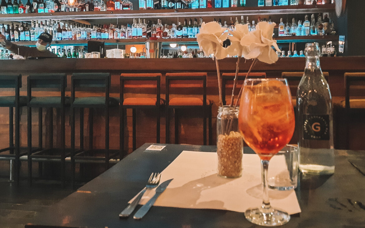 Un Aperol reposando sobre la mesa del Ginger Bar en Mendoza, con el bar encantador y una estantería llena de bebidas al fondo.