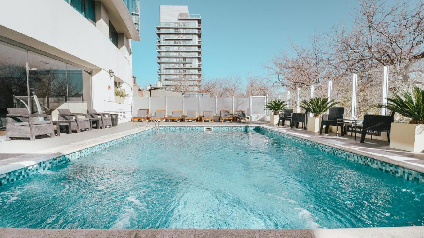 La piscina cristalina del Hotel Diplomático en Mendoza invita al descanso, rodeada de cómodas tumbonas y sillas de terraza bajo un cielo despejado, ofreciendo un oasis de tranquilidad en el entorno urbano.