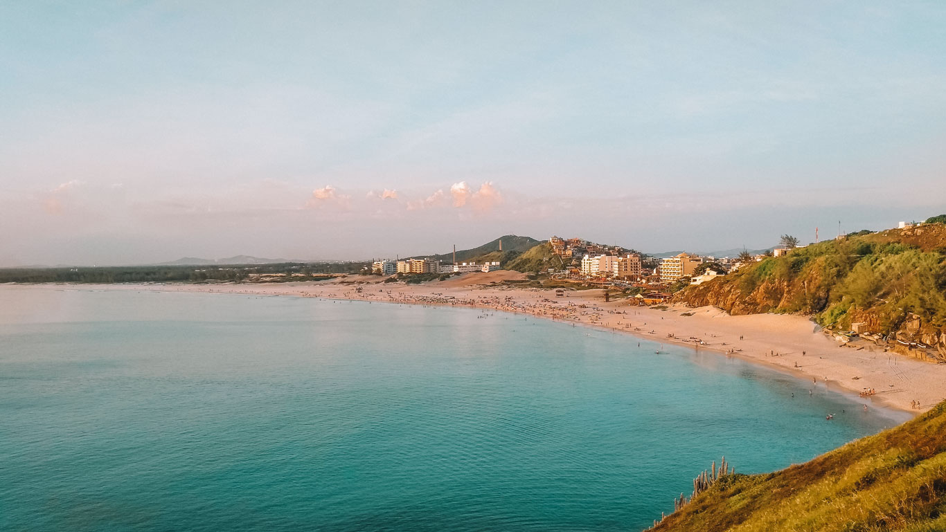 Una amplia vista de la Playa Grande, una de las mejores playas de Arraial do Cabo, captura su extenso arenal y las tranquilas aguas azules, con una densa concentración de visitantes disfrutando del ambiente. Edificaciones y una carretera cercana señalan el desarrollo urbano junto a la playa, todo ello enmarcado por colinas y vegetación costera bajo un cielo suavemente teñido por el atardecer.