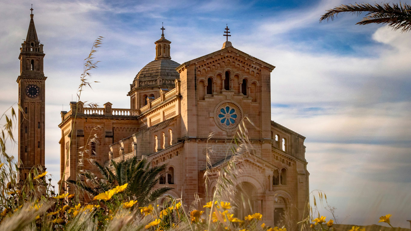 La Basílica de Ta' Pinu en Gozo, Malta, se erige majestuosamente entre las flores silvestres, con su arquitectura ornamentada y sus torres gemelas de campanario que se elevan hacia un cielo azul suave, transmitiendo una sensación de paz y grandeza espiritual.