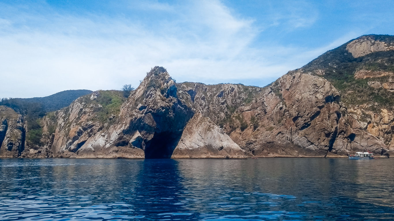 Una embarcación turística se desplaza cerca de la imponente entrada de la Gruta Azul en Arraial do Cabo, con majestuosas formaciones rocosas rodeadas de aguas tranquilas y azules bajo un cielo parcialmente nublado.