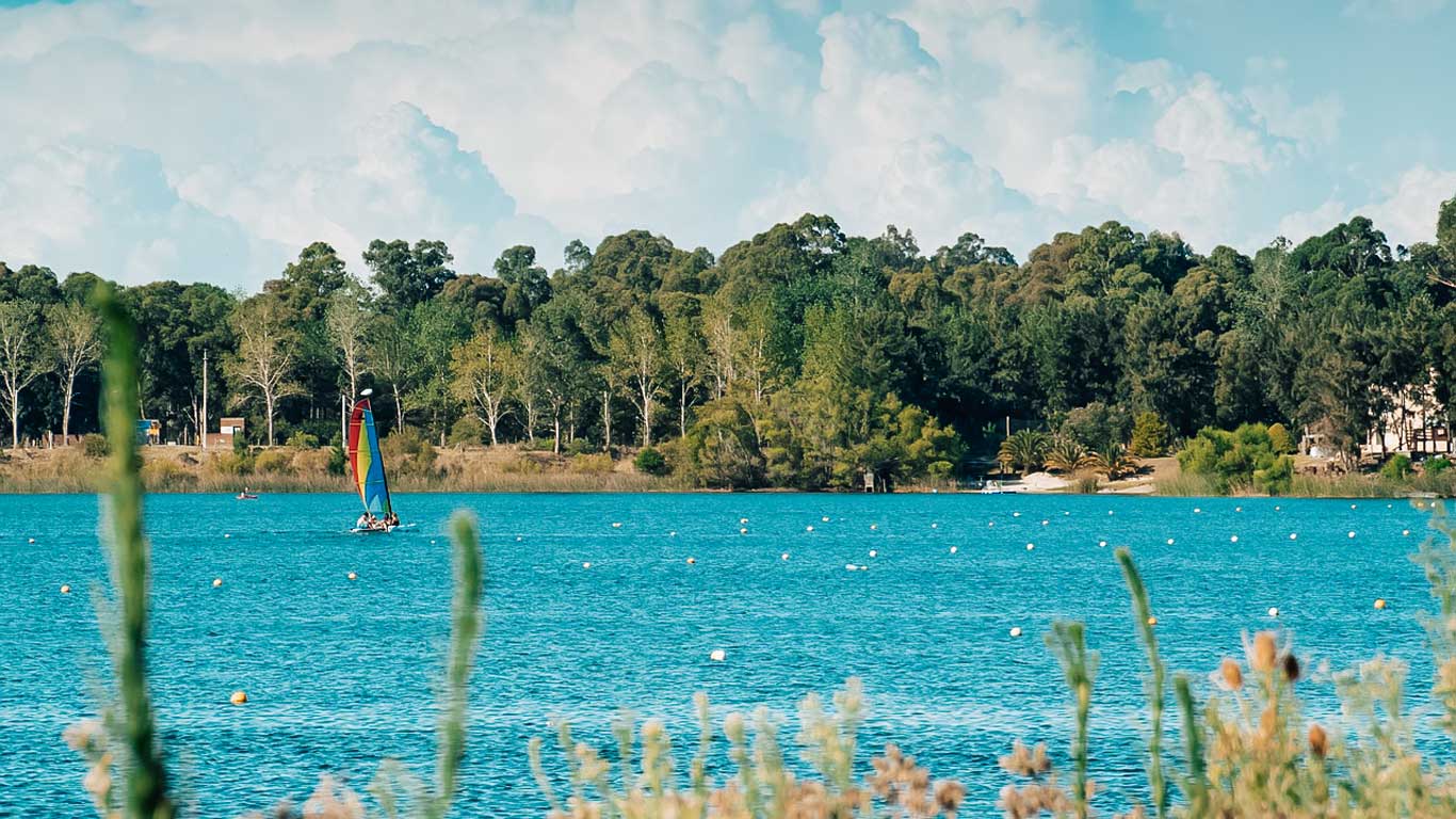 Vista panorámica de la Laguna del Sauce en Punta del Este, con aguas azul turquesa contrastando con la densa vegetación verde al fondo. En el centro de la laguna, una persona practica windsurf con una vela colorida, añadiendo un toque de colores vivos a la escena tranquila.