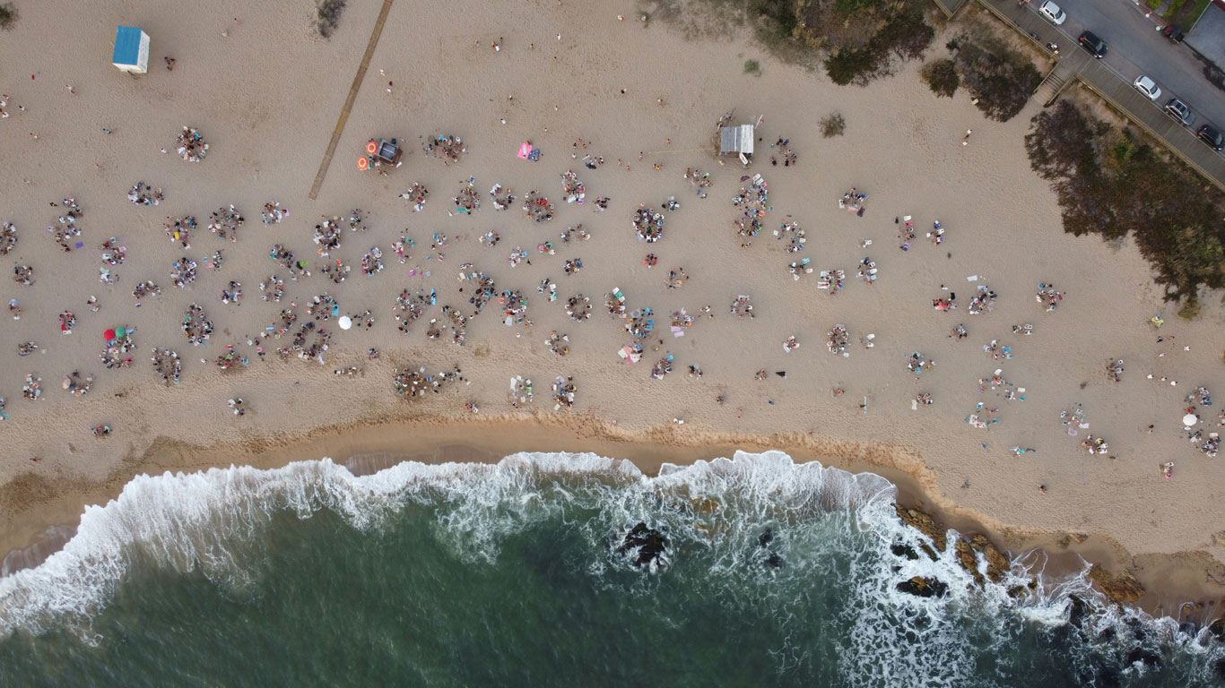 Vista aérea de una escena animada de playa con personas reunidas en la arena, sombrillas y toallas de playa, junto a olas rompiendo en la costa, con una carretera y coches visibles al fondo.
