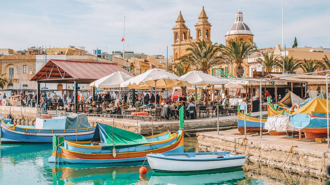 Escena animada del puerto de Marsaxlokk en Malta, con los tradicionales barcos de pesca malteses, conocidos como Luzzus, pintados vivamente y flotando en primer plano, y un bullicioso mercado junto al agua bajo parasoles, enmarcado por edificios históricos y las agujas de las iglesias.