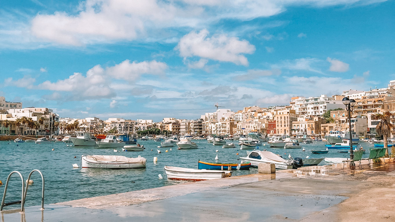 Día luminoso y soleado en Marsaskala, Malta, con una variedad de barcos amarrados en las aguas azules de la bahía, complementados por el pintoresco paseo marítimo adornado con la colorida arquitectura maltesa y palmeras relajantes, evocando un ambiente mediterráneo relajado.