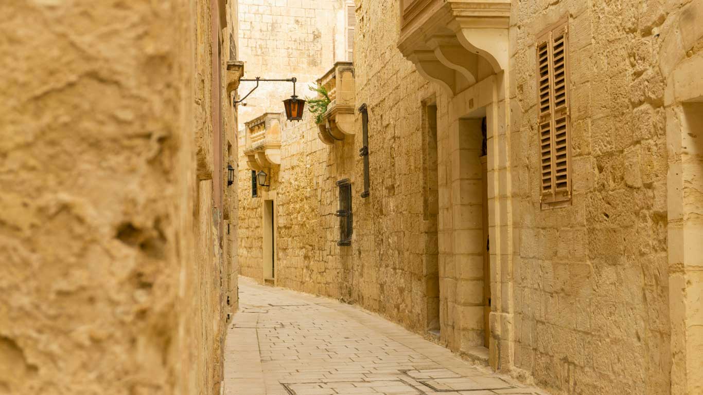 Un estrecho callejón en la antigua ciudad amurallada de Mdina, Malta, flanqueado por tradicionales muros de piedra caliza, adornado con faroles antiguos y evocando una atmósfera atemporal en la "Ciudad Silenciosa".