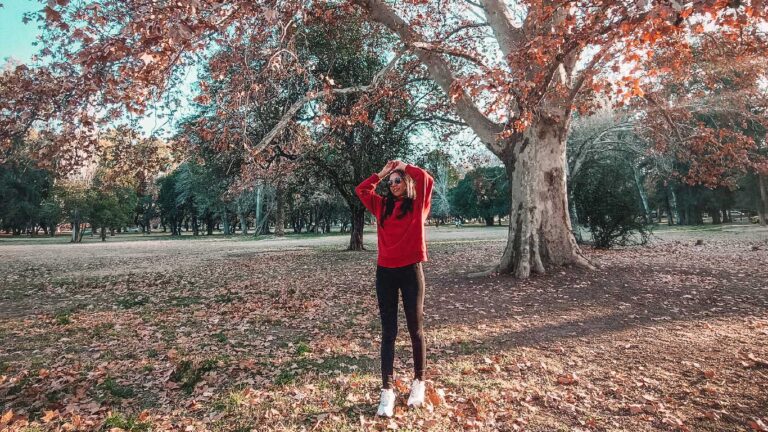 Una mujer con un suéter rojo y pantalones negros, con las manos apoyadas sobre su cabeza en una pose relajada, se encuentra bajo un árbol entre las hojas secas del invierno en el Parque General San Martín en Mendoza.