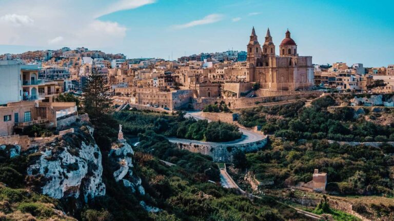 Vista panorámica de una ciudad histórica en Malta, mostrando la arquitectura icónica de piedra color miel y una iglesia prominente con torres gemelas de campanario, situada en lo alto de las colinas con carreteras serpenteantes que conducen a ella, sugiriendo un lugar sereno y pintoresco para alojarse.