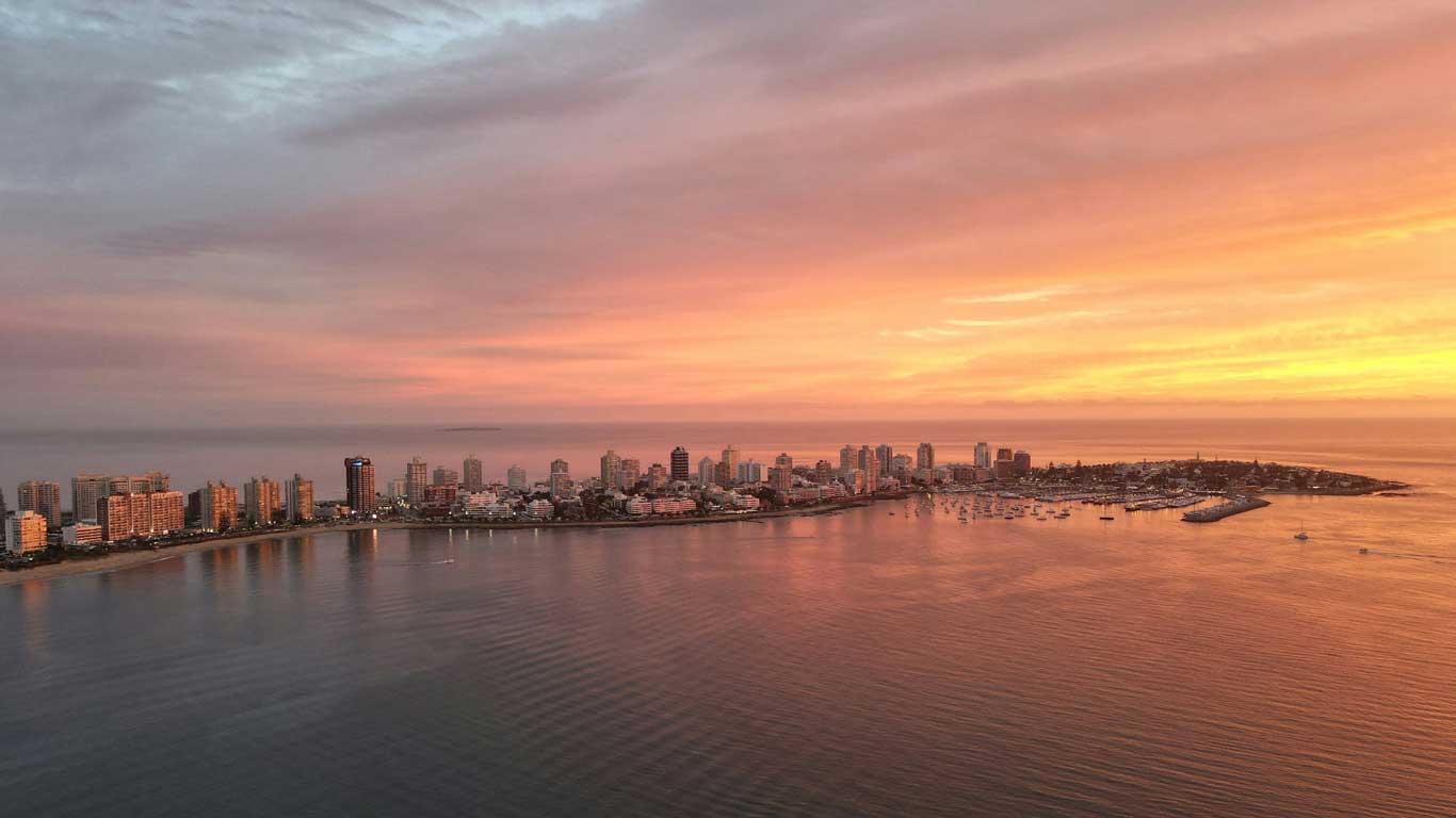 Vista aérea del atardecer tiñendo el cielo de tonos rosados y anaranjados sobre Punta del Este, Uruguay. El paisaje urbano costero está salpicado de rascacielos iluminados que se reflejan en las tranquilas aguas de la bahía, mientras numerosos barcos están anclados en el puerto.