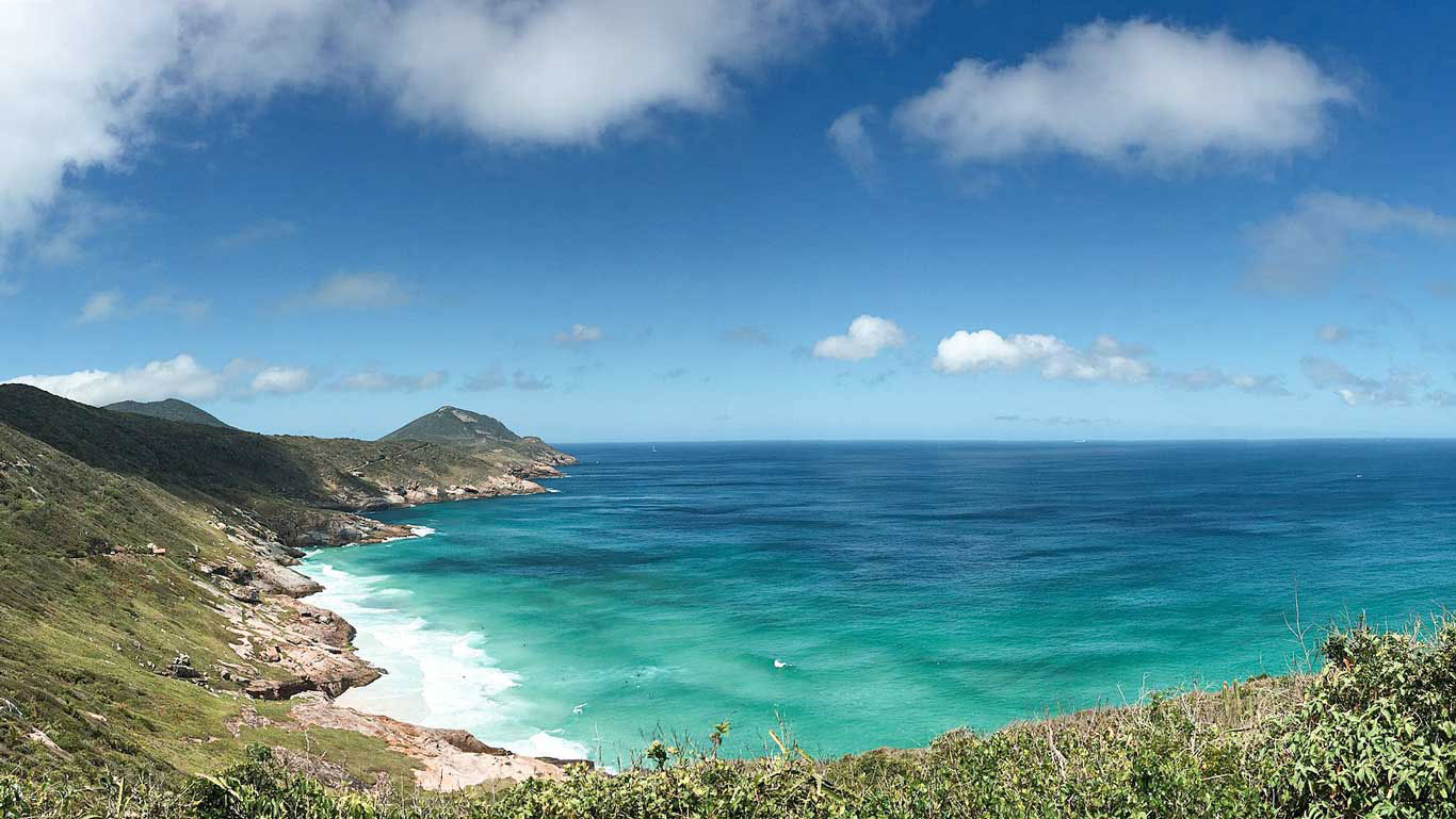 La Playa Brava en Arraial do Cabo se extiende entre dos montañas verdes, revelando su costa rocosa y el mar turquesa que baña su playa apartada. La fotografía panorámica abarca un vasto horizonte marino bajo un cielo azul con nubes esporádicas, enfatizando la serenidad y la belleza natural del paisaje costero.