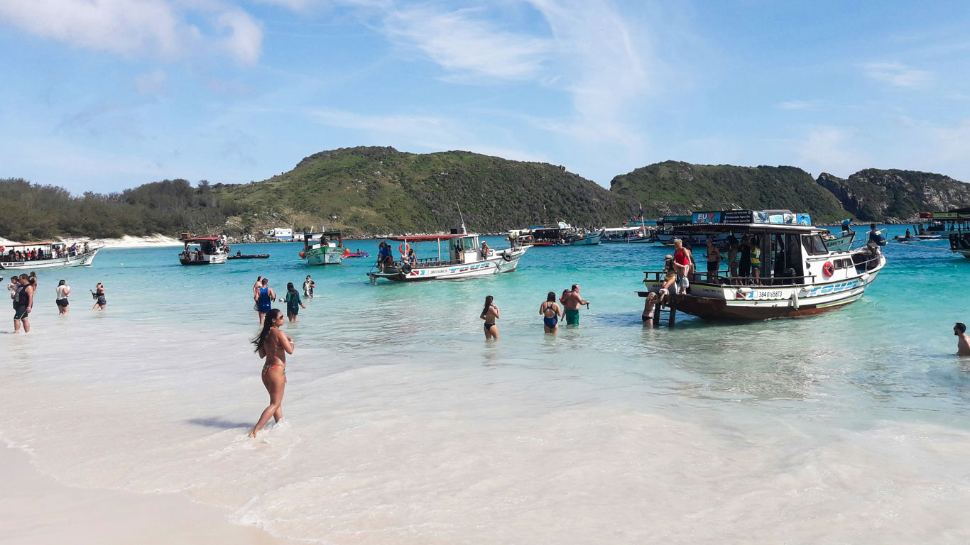 Visitantes y embarcaciones en las cristalinas aguas de la Playa do Farol en Arraial do Cabo, Brasil. Personas bañándose y embarcándose en lanchas bajo un cielo despejado, rodeados por el escenario natural de colinas verdes.