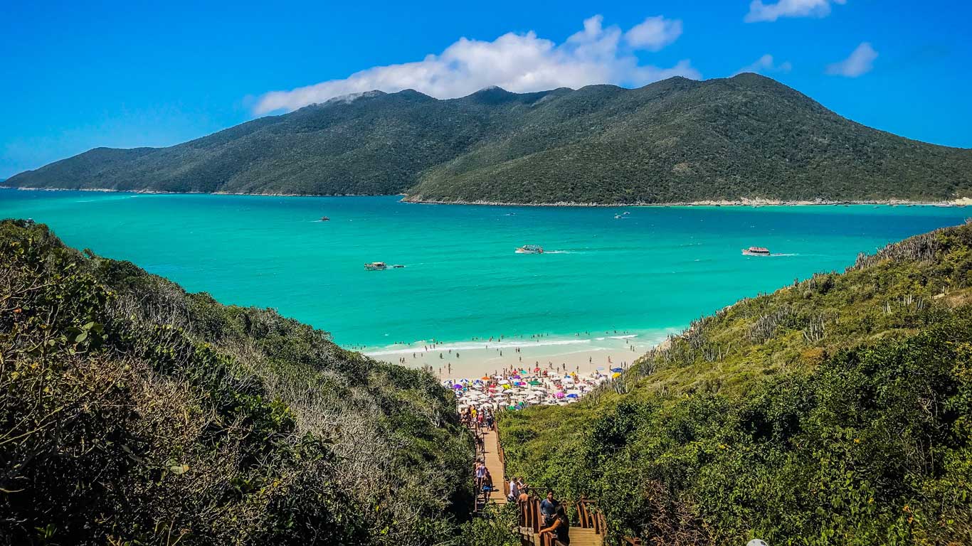 Vista panorámica de las Prainhas do Pontal do Atalaia en Arraial do Cabo, con aguas de color turquesa y una playa llena de visitantes. La imagen muestra una escalera de madera que desciende a través de una vegetación densa hacia la arena blanca, flanqueada por montañas verdes bajo un cielo azul.