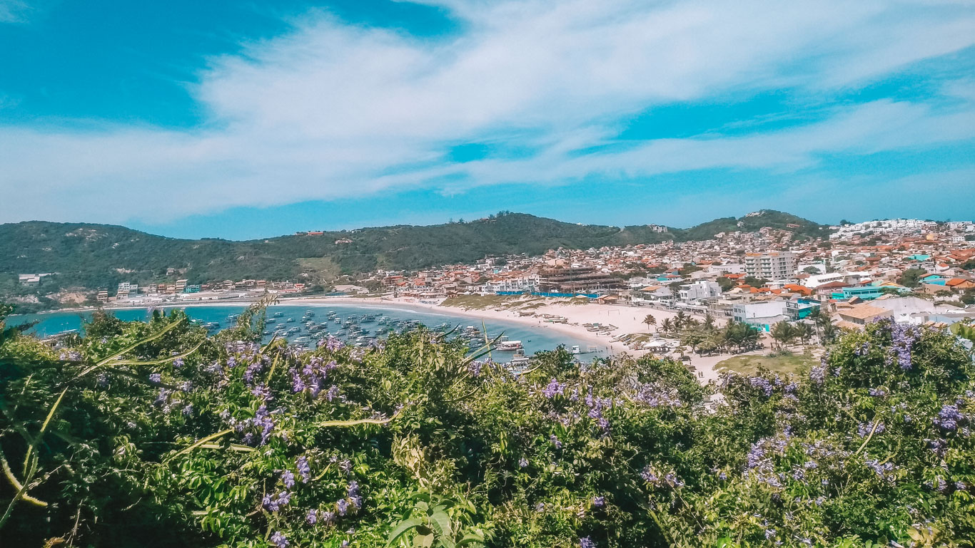 La fotografía muestra la Playa dos Anjos en Arraial do Cabo, rodeada de una ciudad costera. La playa se curva suavemente junto a un mar tranquilo y azul, con embarcaciones dispersas cerca de la orilla. En primer plano, flores silvestres añaden color natural a la escena urbana y playera bajo un cielo azul claro.