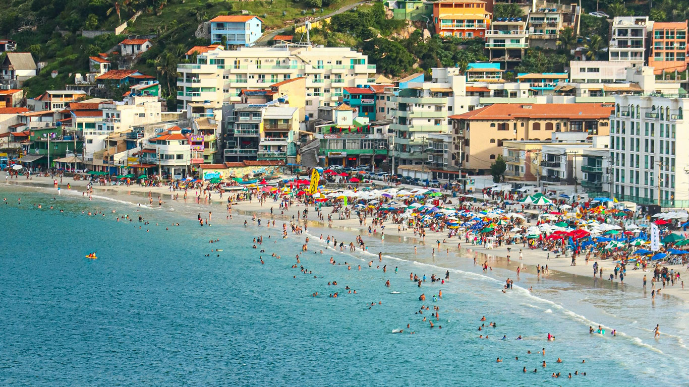 La Prainha en Arraial do Cabo bulle de actividad con multitudes de bañistas y coloridos paraguas a lo largo de su arena blanca. Edificios de varios pisos se alinean justo en el borde de la playa, ofreciendo un contraste vívido entre el ocio de playa y la vida urbana, todo ello enmarcado por las aguas azules del mar y un cielo claro.