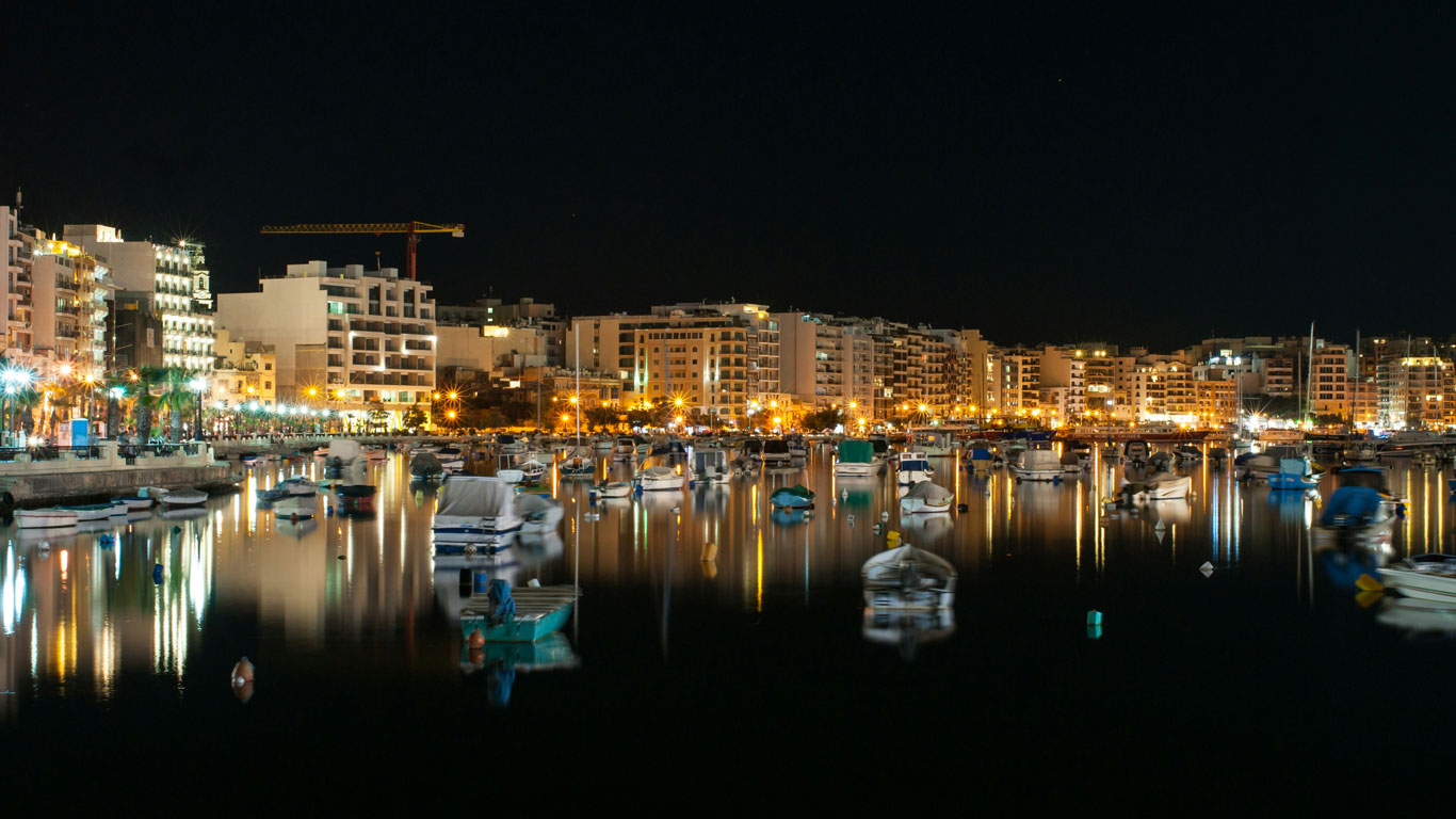 Vista nocturna de Sliema en Malta, con las luces de la ciudad brillando sobre el mar en calma, salpicado de barcos amarrados creando una escena serena y a la vez vibrante en el paseo marítimo.