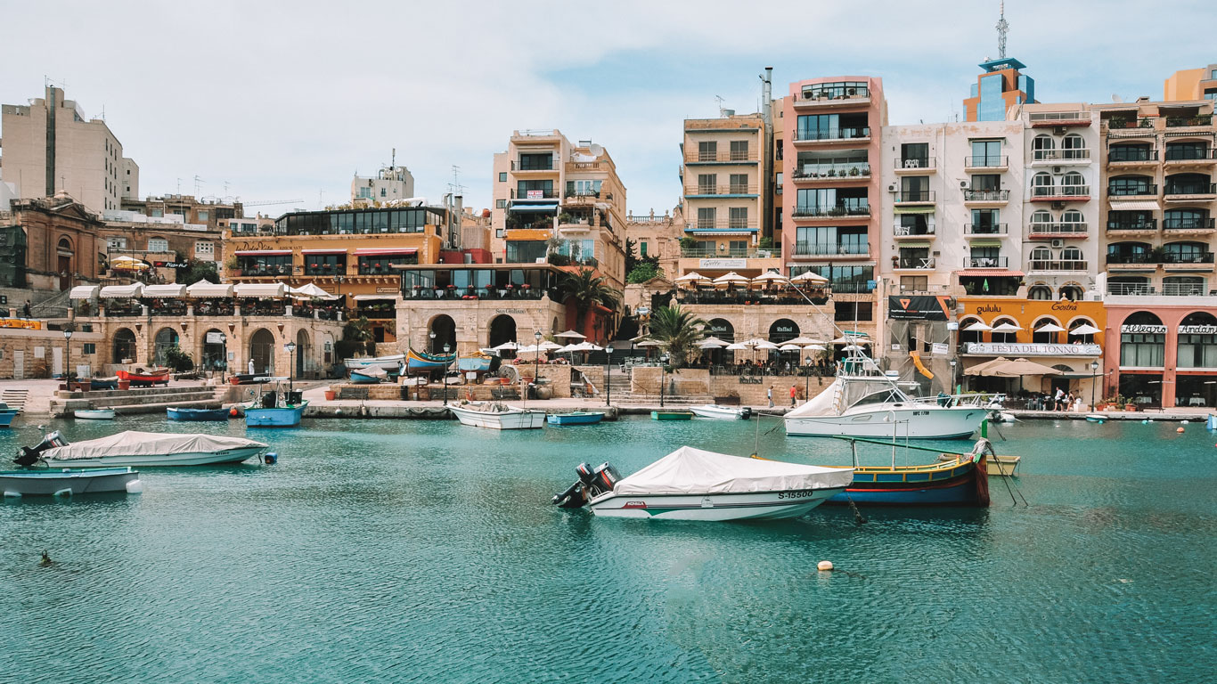 Escena colorida junto al agua en St. Julian's en Malta, con barcos flotando en las aguas color aguamarina y un telón de fondo de restaurantes pintorescos y edificios de varios pisos que capturan la vivaz vida costera.